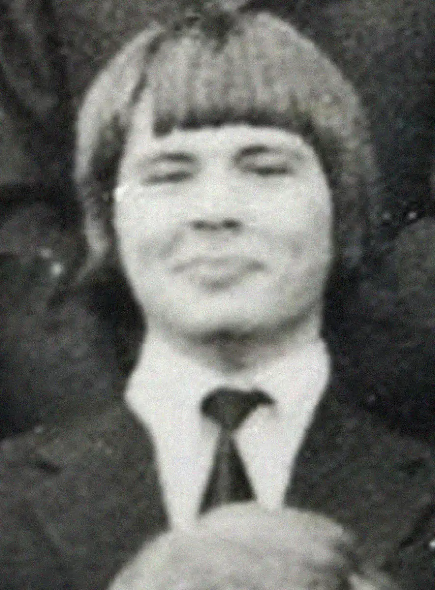 A black and white photo of a young person with straight, chin-length hair and bangs, wearing a suit jacket, dress shirt, and tie, smiling slightly at the camera.