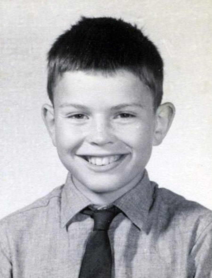 A young boy with short dark hair smiles at the camera. He is wearing a light-colored shirt with a collar and a dark tie. The photo is black and white with a plain background.