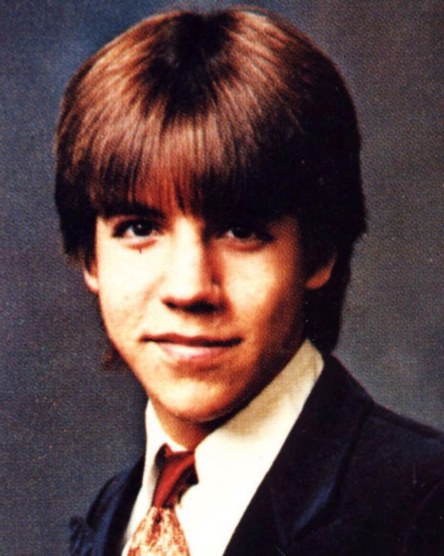 A young person with straight brown hair in a bowl cut, wearing a dark suit jacket, white shirt, and patterned tie, smiling slightly at the camera against a plain background.