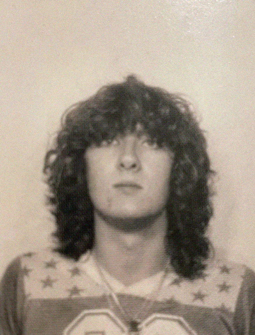 Black and white photo of a young person with curly, shoulder-length hair, wearing a shirt with stars on the shoulders and the number "82" visible on the front. The individual is facing forward with a neutral expression.