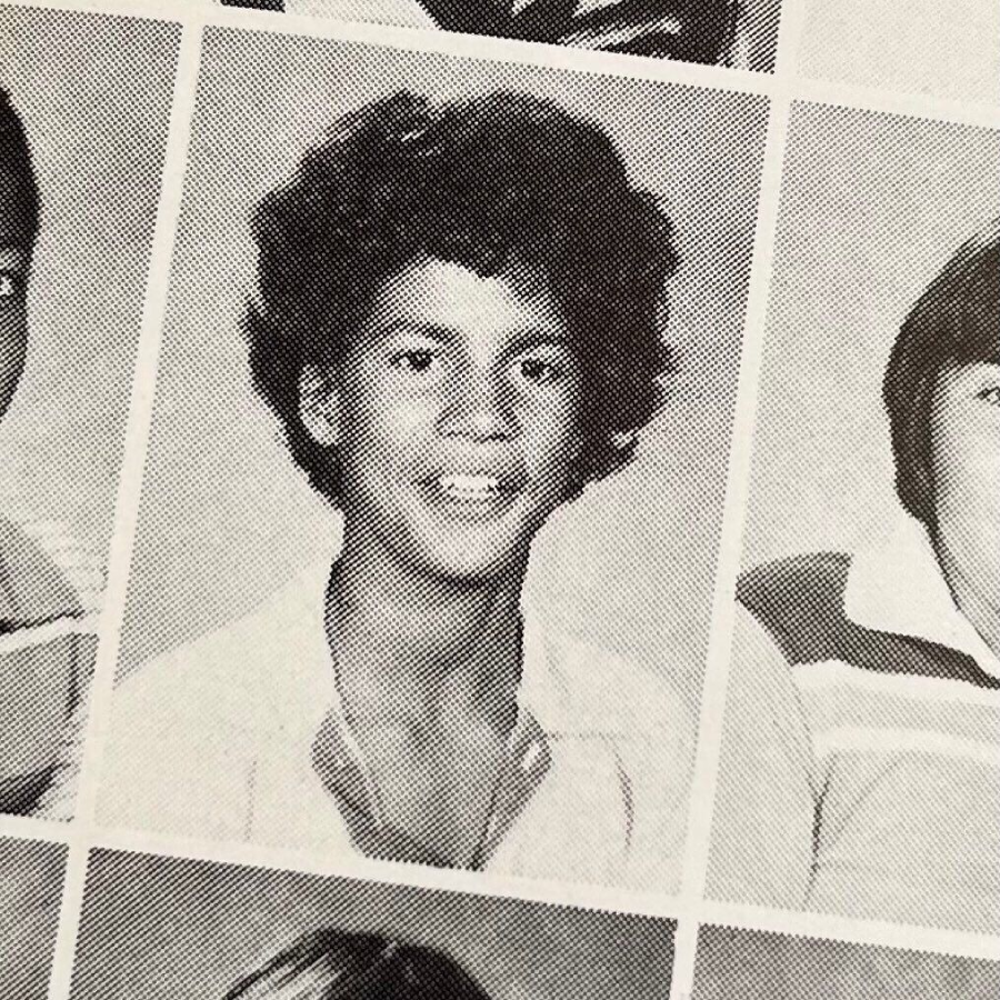Black and white yearbook photo of a young person with curly hair, smiling, wearing an open-collared shirt. The image is part of a grid with other student portraits visible around it.