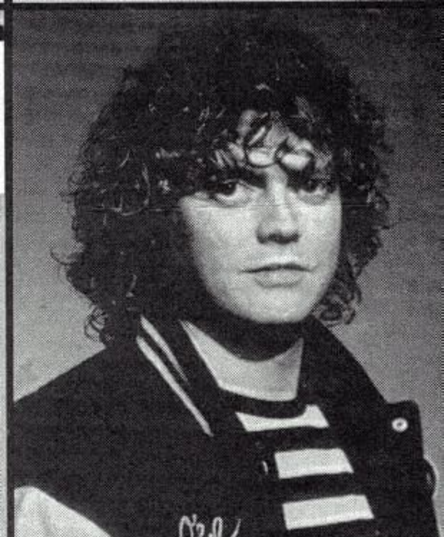 Black and white portrait of a person with curly hair, wearing a striped shirt and a jacket with a letter on it, looking directly at the camera with a neutral expression.