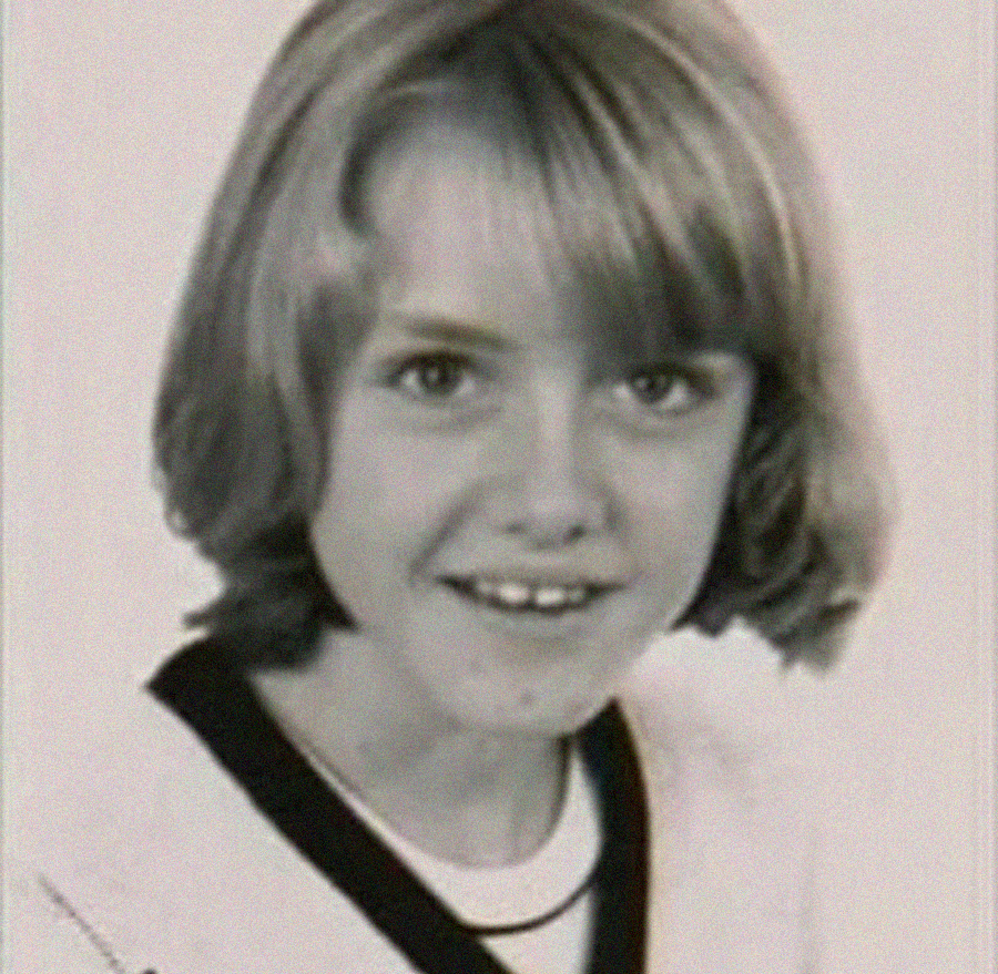 A black-and-white portrait of a young girl with light-colored, shoulder-length hair and bangs, smiling at the camera while wearing a sweater with a dark collar.