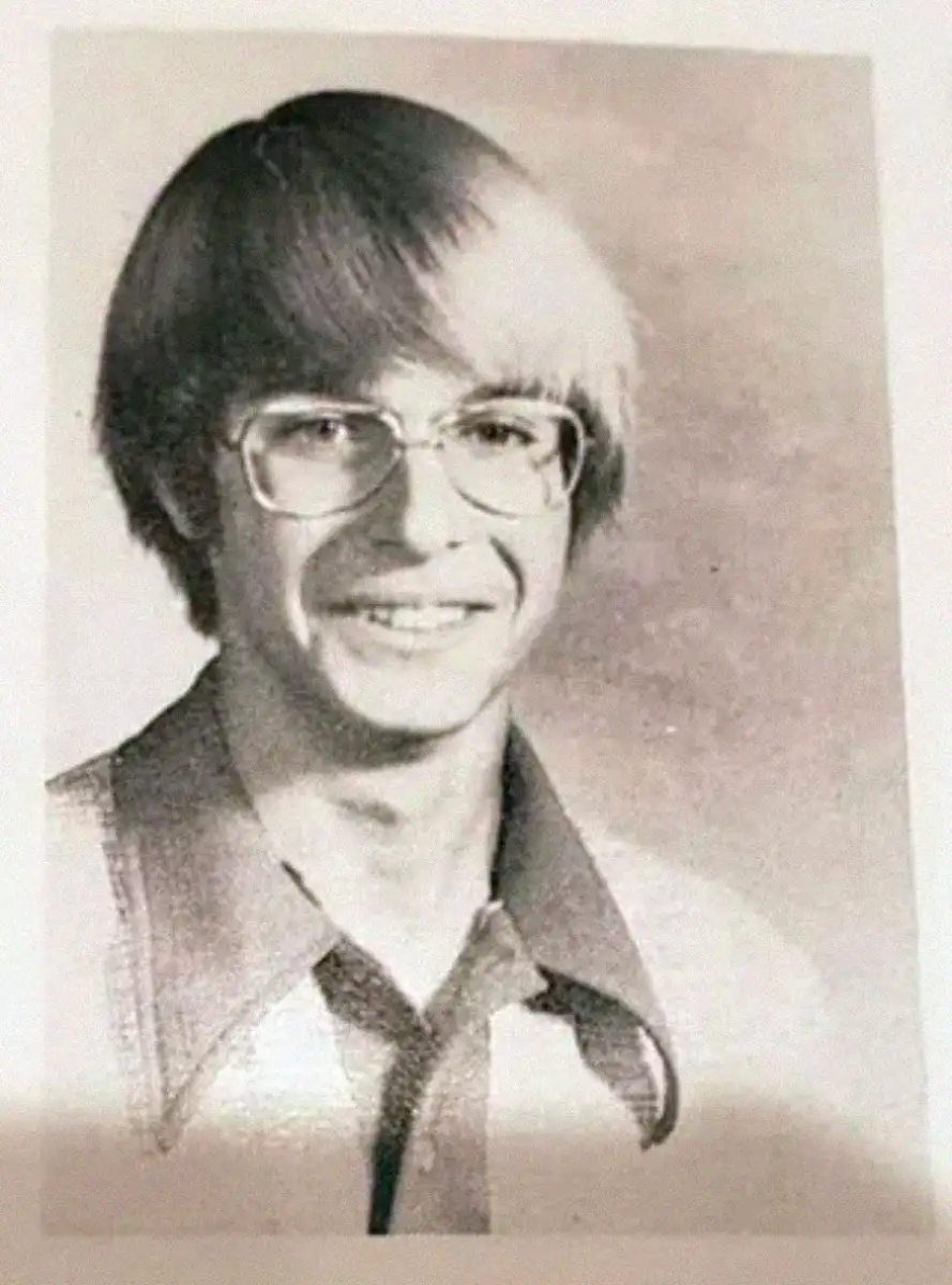 Black-and-white portrait of a young man with straight, light hair and glasses, smiling. He is wearing a collared shirt with wide lapels and a tie. The background is plain and softly blurred.