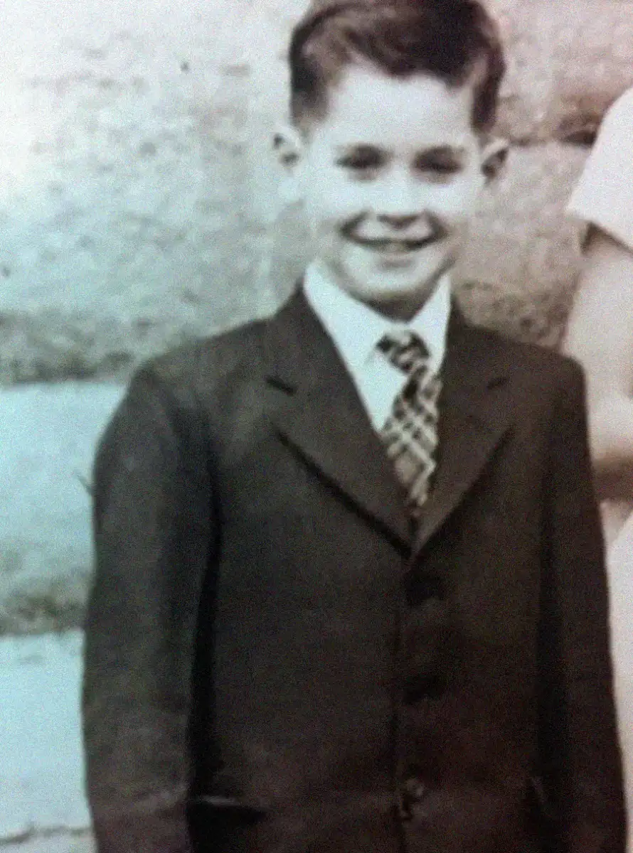 A black-and-white photo of a smiling young boy wearing a suit and patterned tie, standing in front of a stone wall. Only part of another person is visible on the right side of the image.