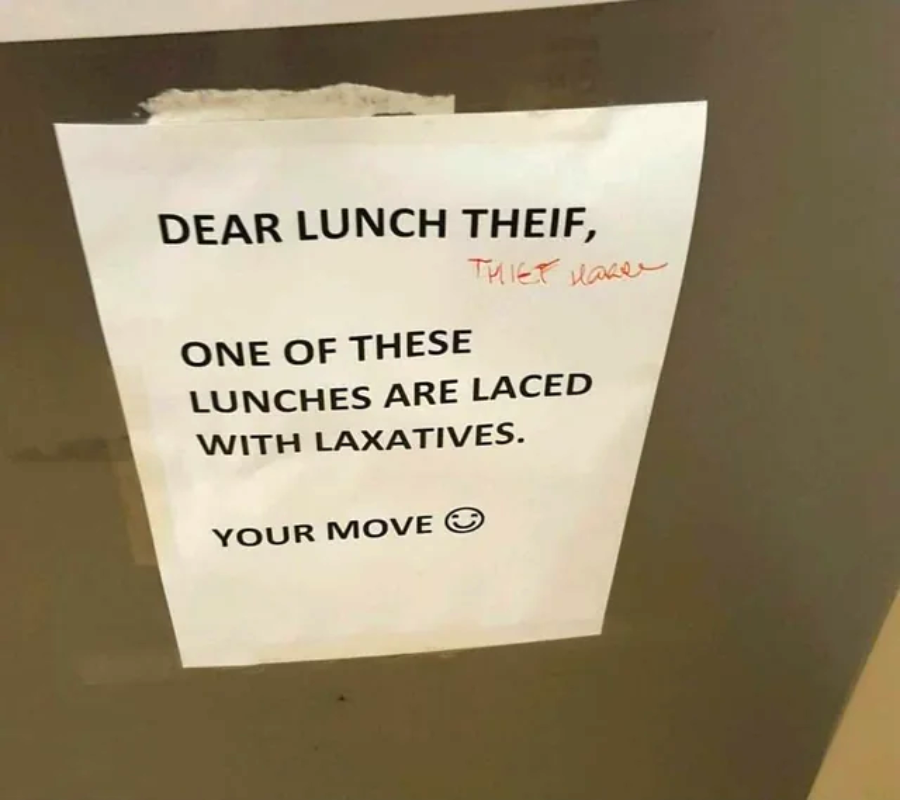 A printed sign taped to a surface reads: "DEAR LUNCH THEIF, ONE OF THESE LUNCHES ARE LACED WITH LAXATIVES. YOUR MOVE ;)" with “THEIF” corrected to “THIEF” in red pen.