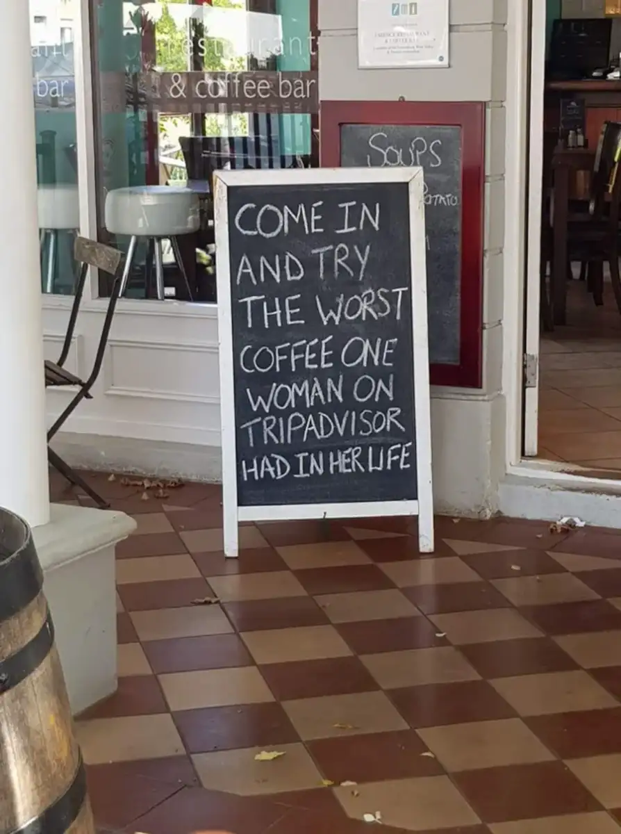 A sidewalk sign outside a café reads, "Come in and try the worst coffee one woman on TripAdvisor had in her life." The sign sits on a tile floor near the café entrance.
