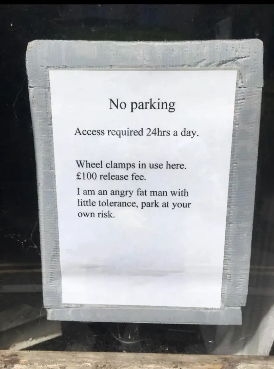 A printed, taped sign reads: "No parking. Access required 24hrs a day. Wheel clamps in use here. £100 release fee. I am an angry fat man with little tolerance, park at your own risk.