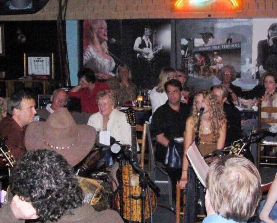 A crowded music venue with people seated at tables, listening to musicians performing onstage. Guitars and music stands are visible, and large posters of musicians decorate the wall behind the audience.