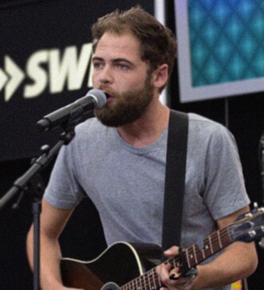 A bearded man wearing a gray t-shirt sings into a microphone while playing an acoustic guitar on stage. A blurred sign and digital screen are visible in the background.