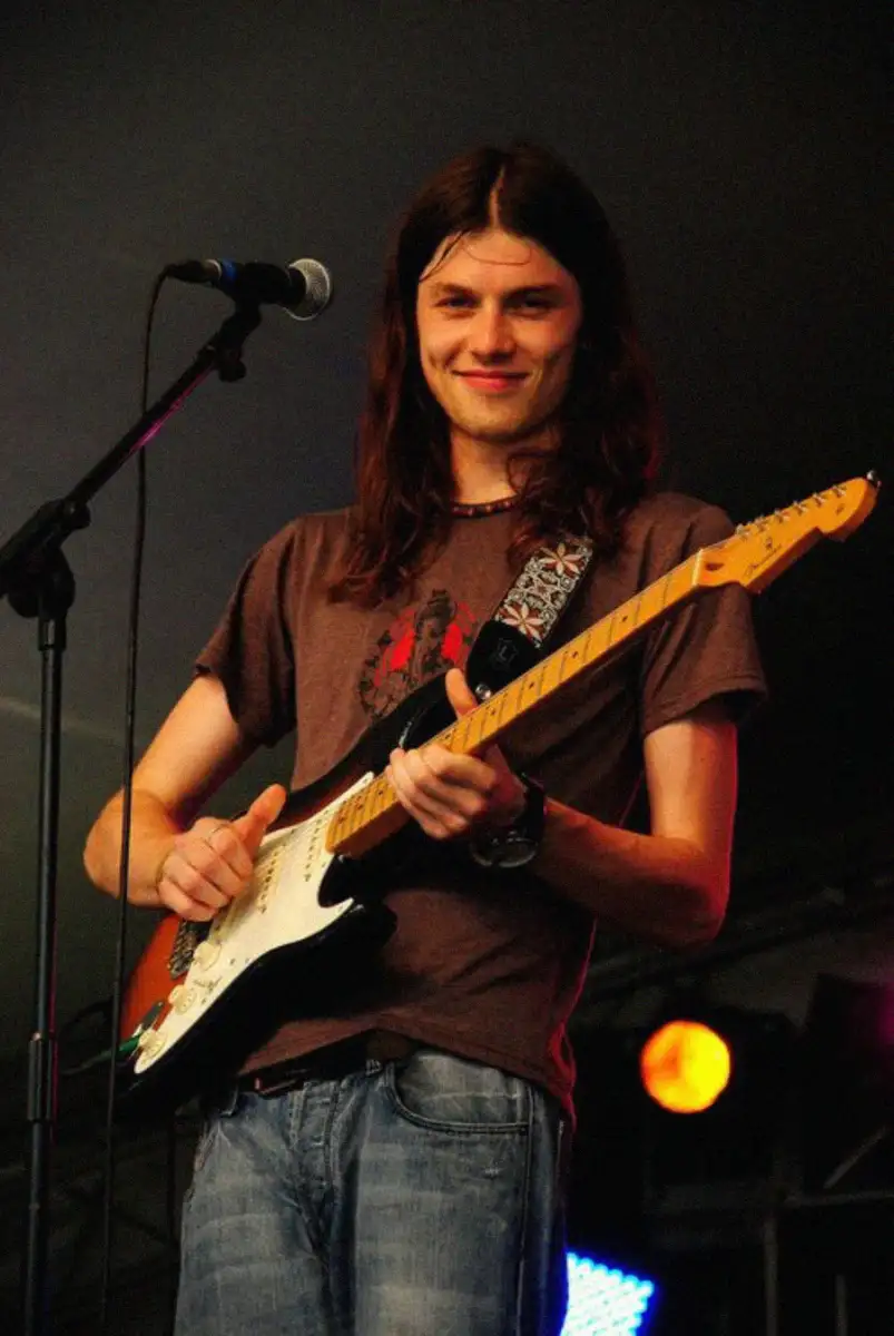 A young man with long brown hair plays an electric guitar on stage, smiling at the camera. He wears a brown T-shirt, jeans, and a beaded necklace. A microphone and colorful stage lights are visible in the background.