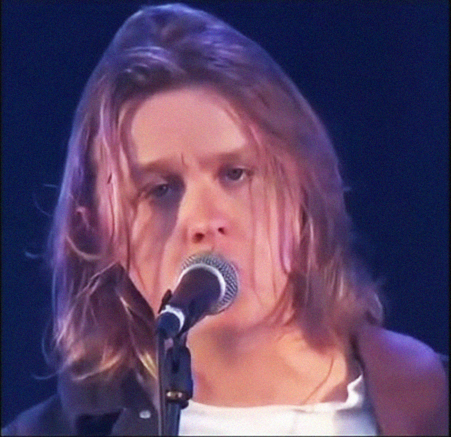 A person with long, light brown hair is singing into a microphone on stage, wearing a dark shirt over a white top, with a focused expression.