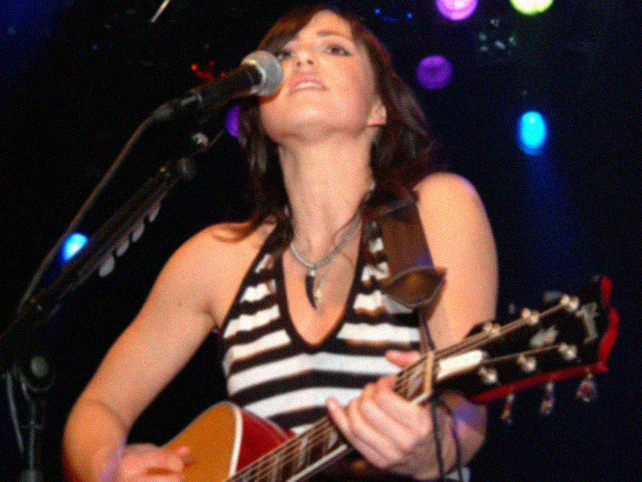 A woman in a sleeveless black-and-white striped top plays an acoustic guitar and sings into a microphone on stage, with colorful lights shining in the background.