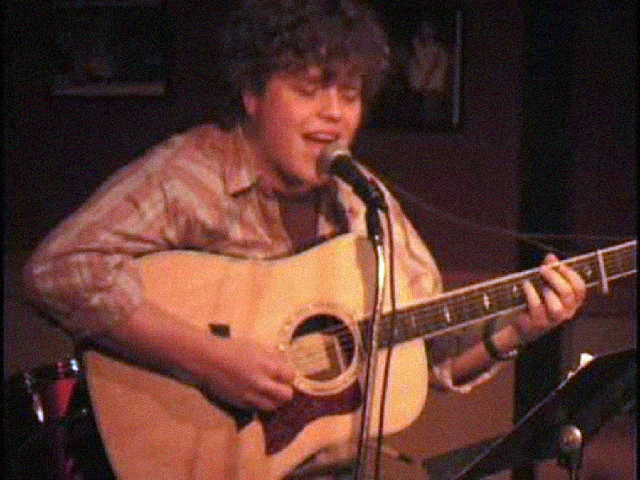 A person with curly hair plays an acoustic guitar and sings into a microphone on stage, wearing a plaid shirt. The background is dimly lit with framed photos on the wall.