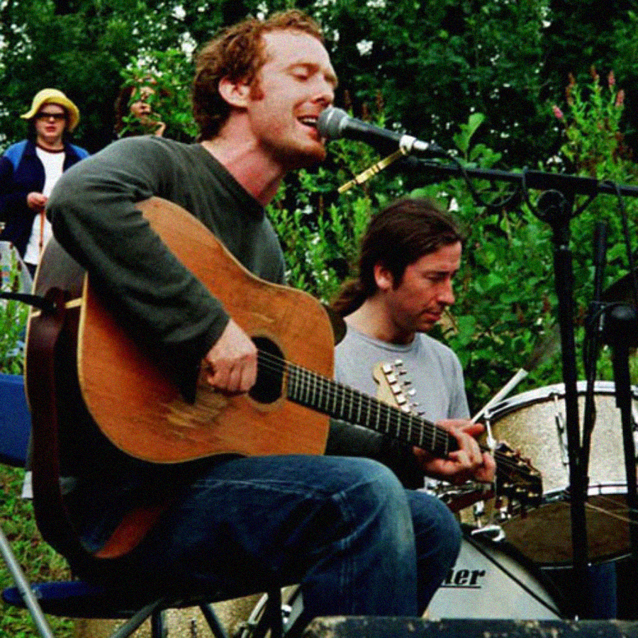 A man sits outdoors playing an acoustic guitar and singing into a microphone, with another musician and a drum set behind him. Trees and people are visible in the background.