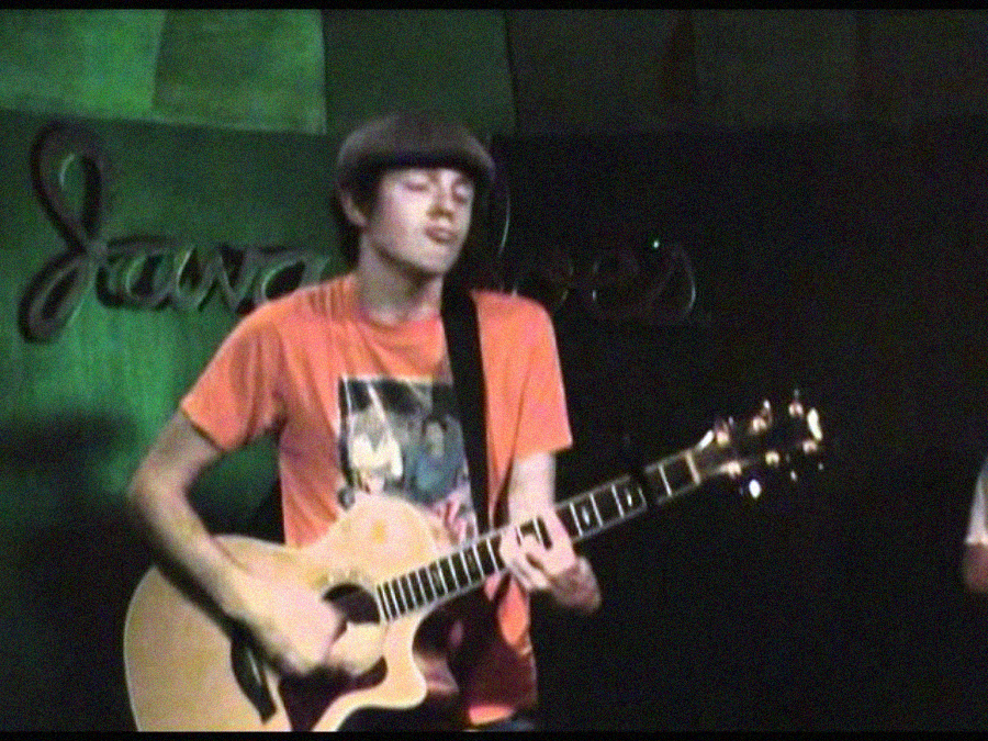 A person in an orange shirt and flat cap plays an acoustic guitar on stage, with partially visible text and green lighting in the background.