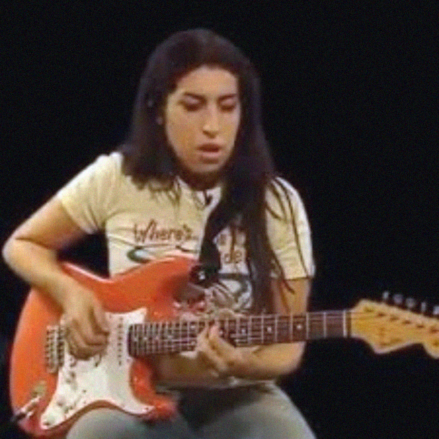 A woman with long dark hair plays a red electric guitar while wearing a light-colored T-shirt, seated against a black background.