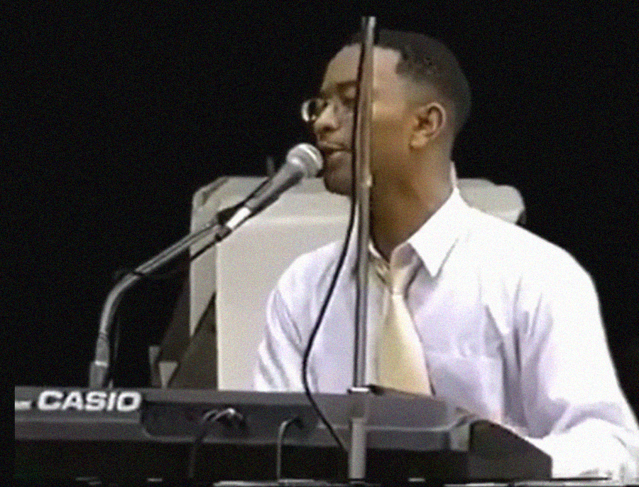 A man in glasses and a white shirt with a light tie plays a Casio keyboard and sings into a microphone on stage.
