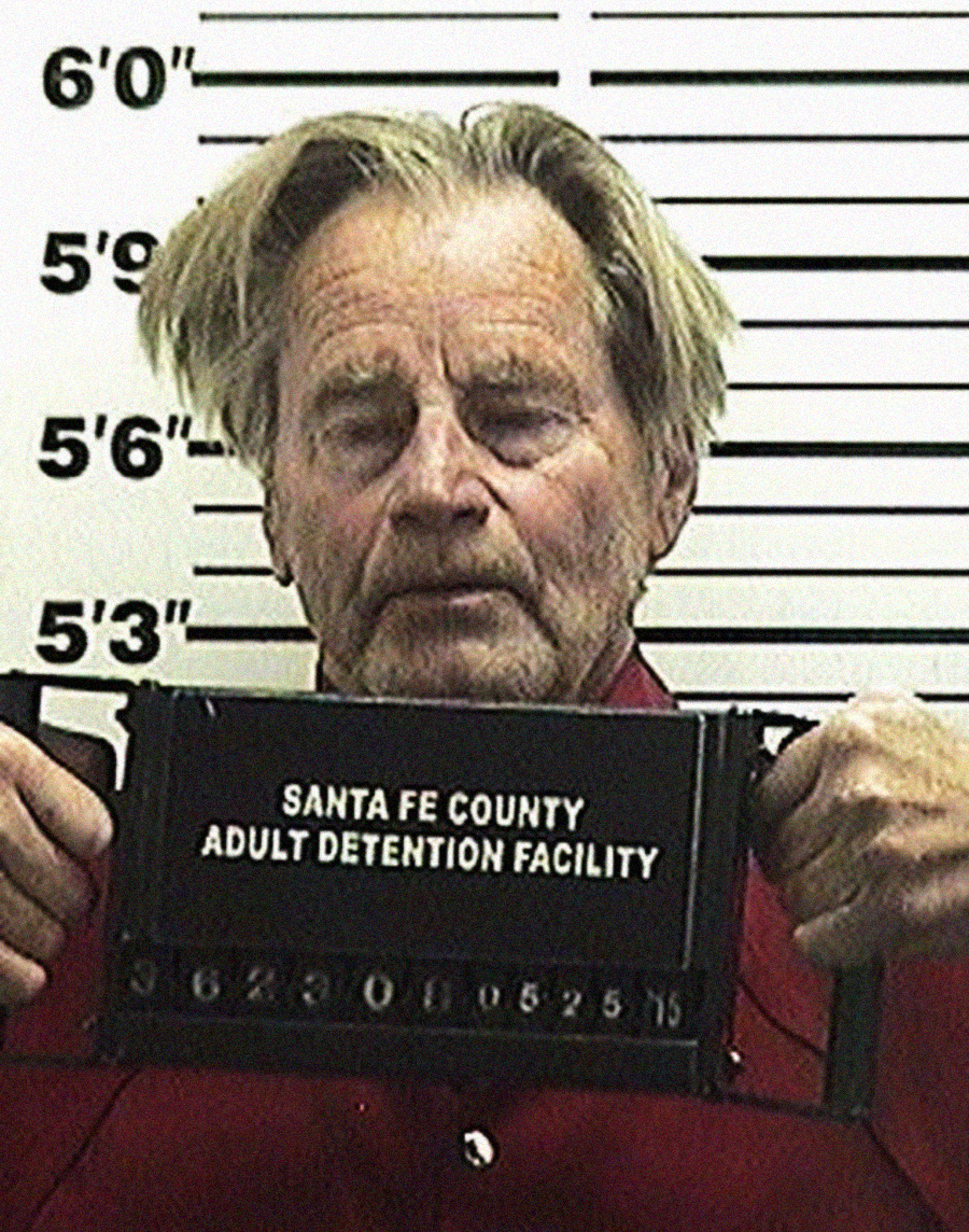 A man with gray hair and a beard stands against a height chart holding a sign that reads "Santa Fe County Adult Detention Facility." His eyes are partially closed and he is wearing a red shirt.