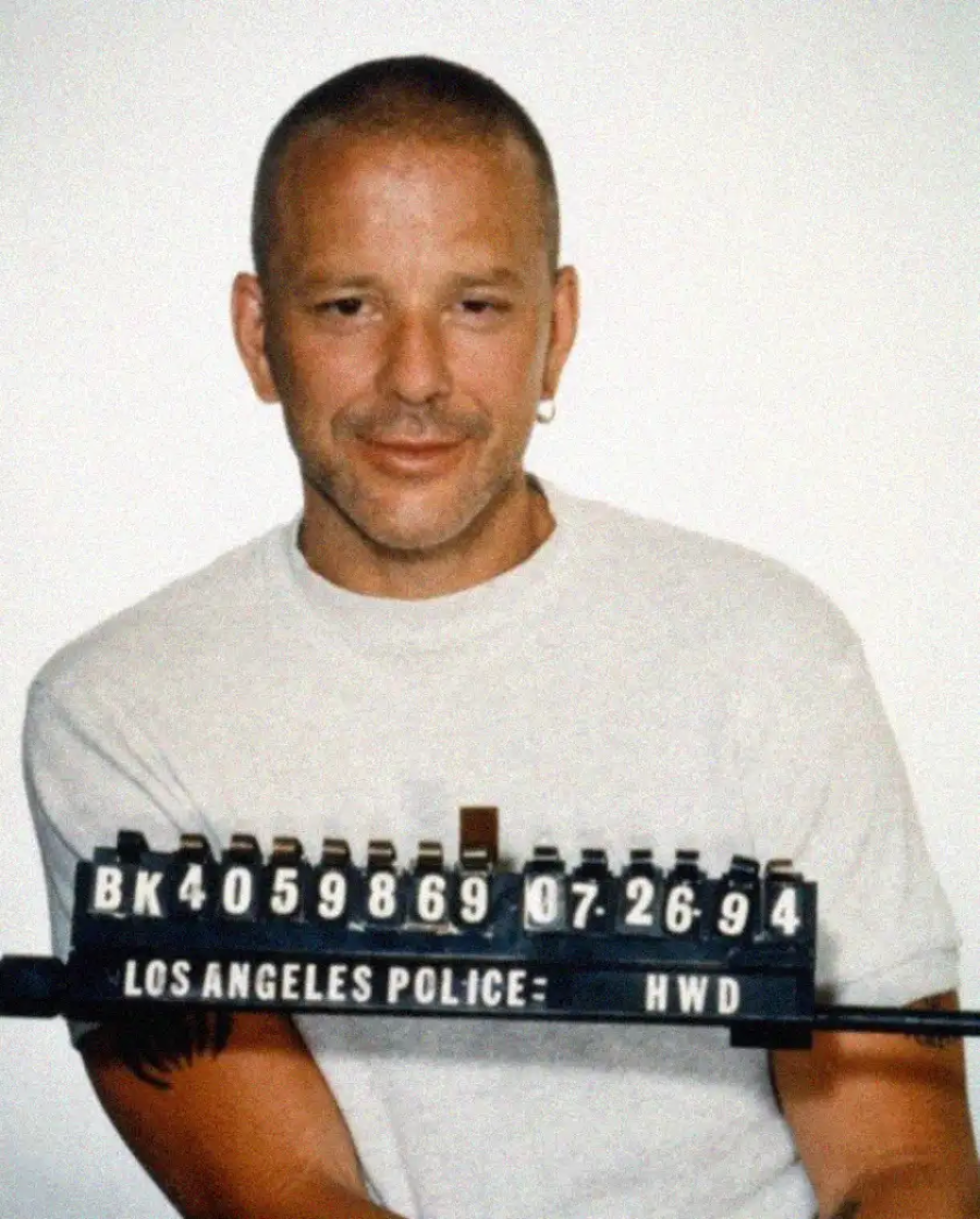 A man with short hair, wearing a white T-shirt and a hoop earring, poses for a police mugshot. He is smiling slightly, and the sign in front reads "LOS ANGELES POLICE" with numbers and a date.