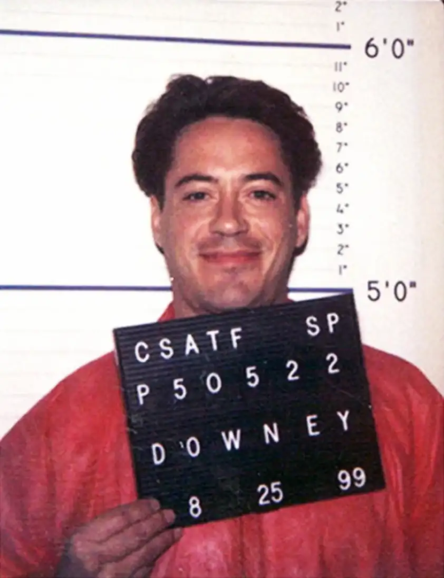 A man in a red shirt smiles while holding a black ID board in front of a height chart, as part of a mugshot. The board displays numbers, letters, and the name "Downey.