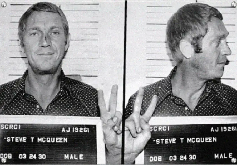 Black-and-white mugshot of a man in a polka-dotted shirt holding up a peace sign with his right hand. The front and side profile are shown. The sign includes the name "Steve T McQueen.