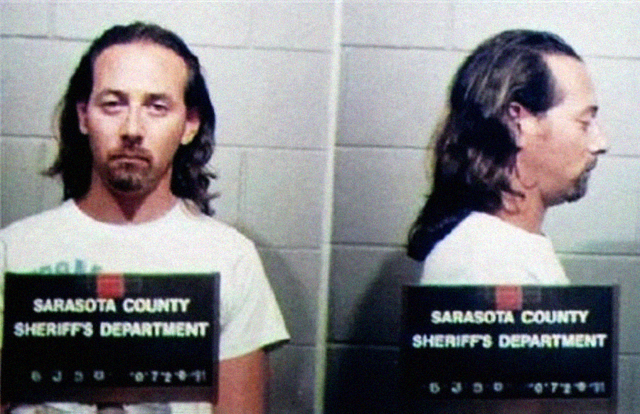 A man with long hair wearing a white t-shirt is shown in front and side mugshots holding a sign that reads "Sarasota County Sheriff's Department." The background is made of cinder block walls.