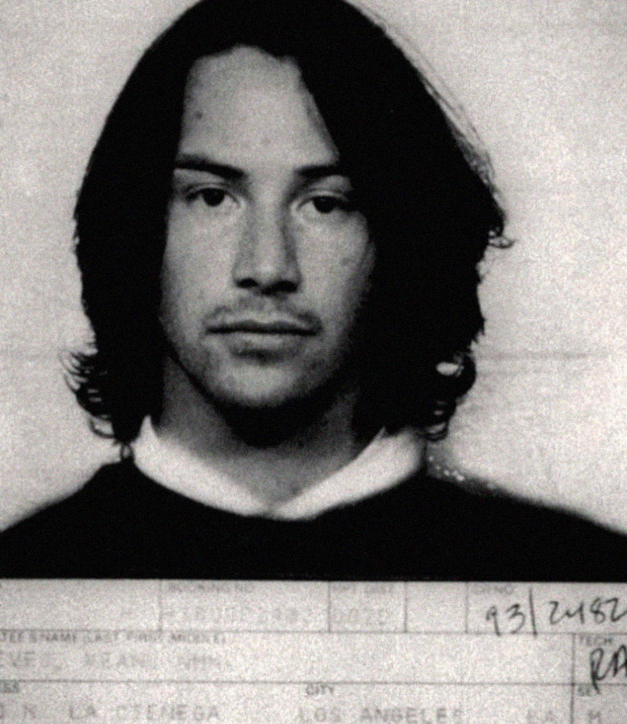 Black and white portrait photograph of a young man with shoulder-length dark hair, wearing a dark sweater over a white collared shirt, with a neutral expression. There is partial text visible at the bottom of the image.