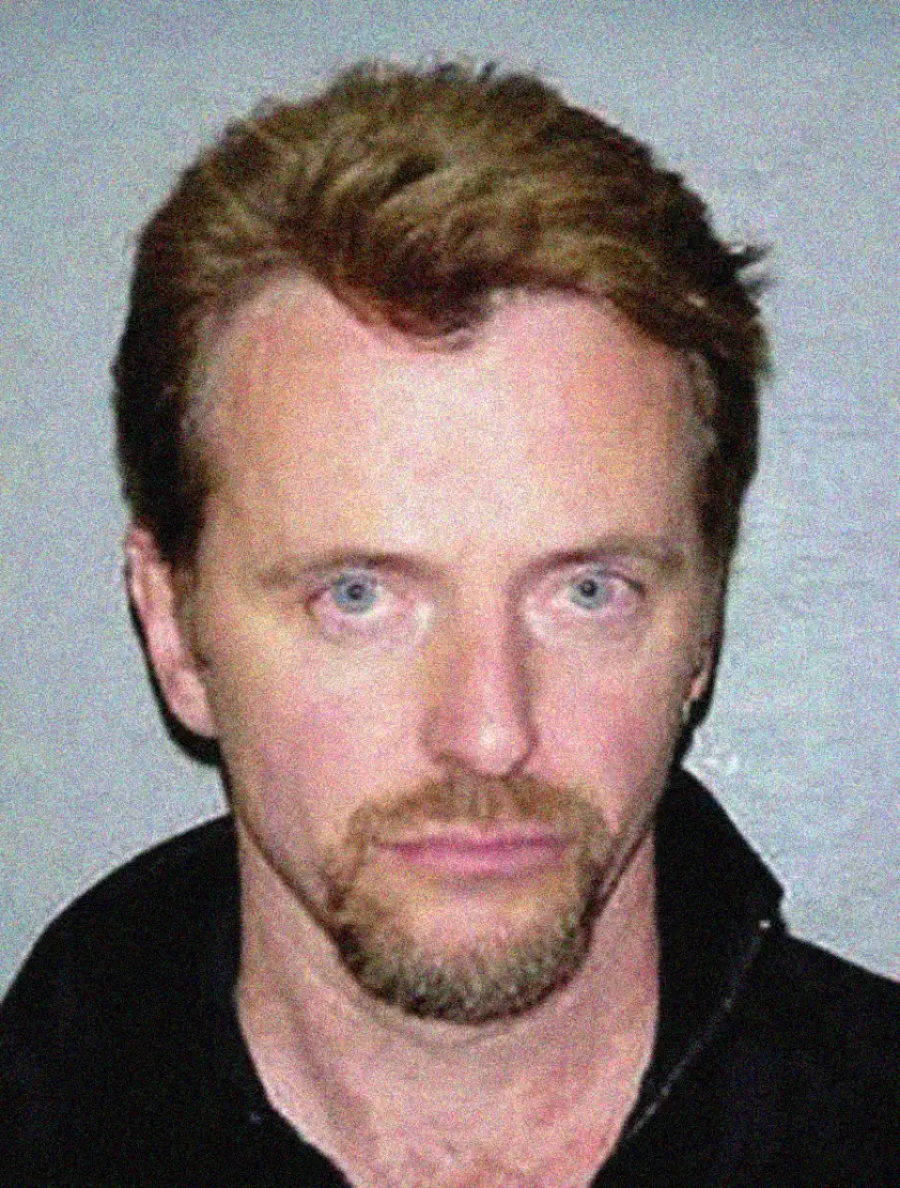 A man with short light brown hair, a mustache, and a goatee is wearing a dark jacket and looking directly at the camera against a plain gray background.