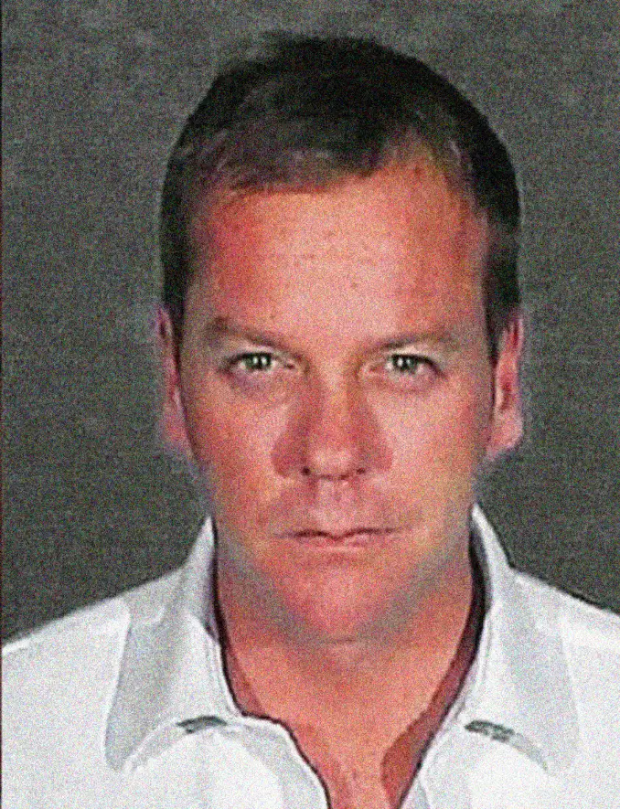 A man with short hair wearing a white collared shirt looks directly at the camera against a plain, dark background. The image has a serious tone and appears to be a mugshot.