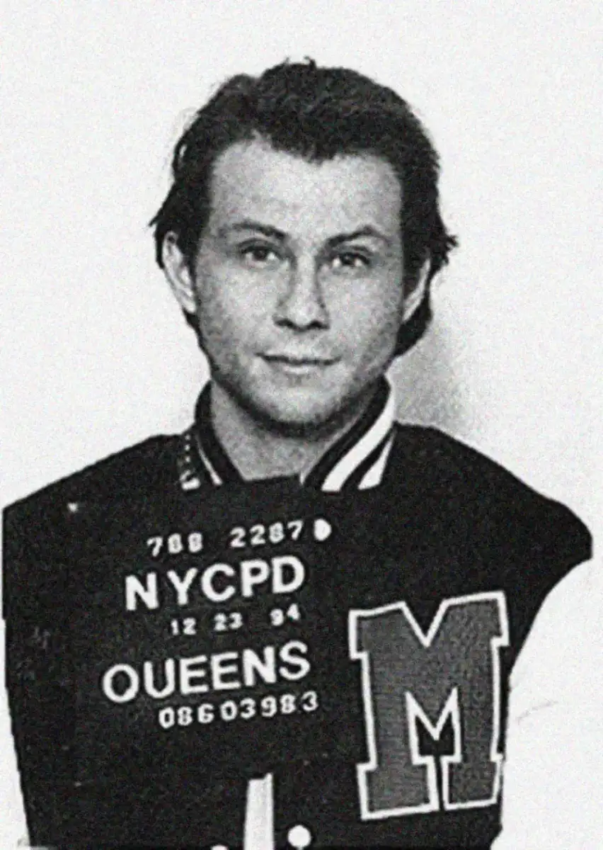 Black and white mugshot of a young man with dark hair, wearing a varsity jacket with a large “M” on it, holding a sign that reads "NYCPD Queens" and showing identification numbers.
