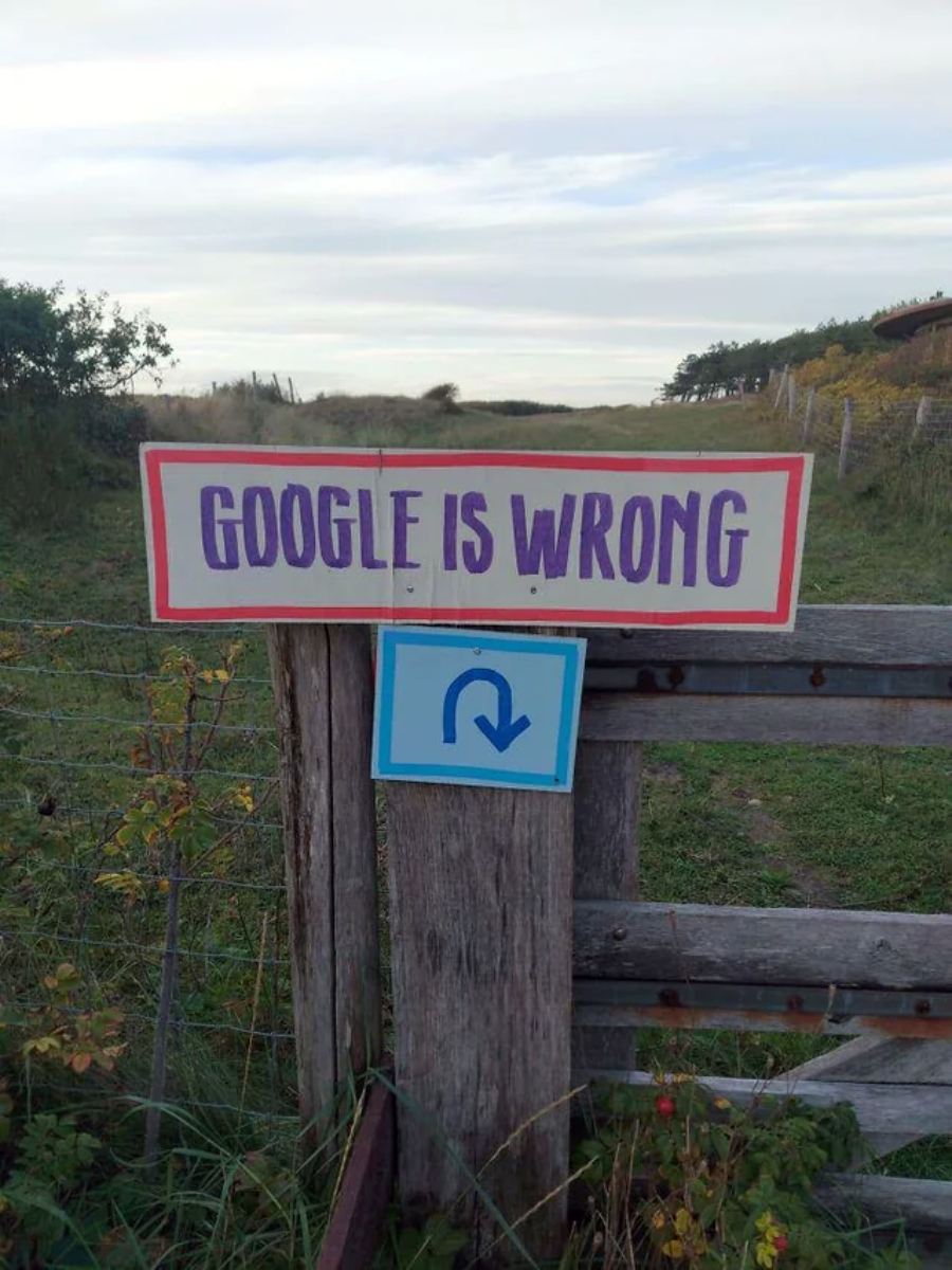 A sign on a wooden fence reads "GOOGLE IS WRONG" with a smaller sign below showing a U-turn arrow, in a rural setting with grassy fields and a cloudy sky in the background.