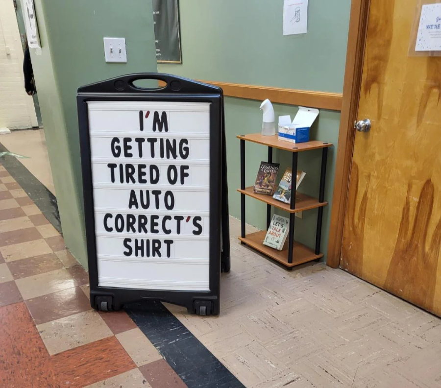 A sign in a hallway reads, "I'M GETTING TIRED OF AUTO CORRECT'S SHIRT." A small shelf with tissues, hand sanitizer, and books is nearby against a green and tan wall.