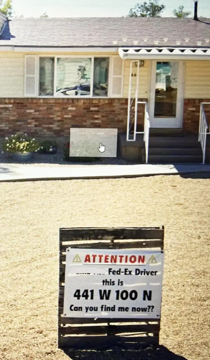 A sign in front of a house reads, "ATTENTION Fed-Ex Driver this is 441 W 100 N Can you find me now?" The house has tan siding, brick trim, a porch, and gravel yard.