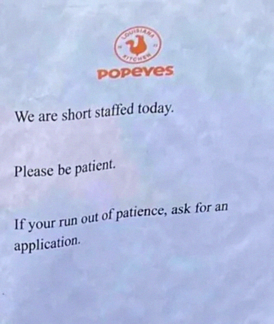 A printed sign with the Popeyes logo reads: "We are short staffed today. Please be patient. If you run out of patience, ask for an application.