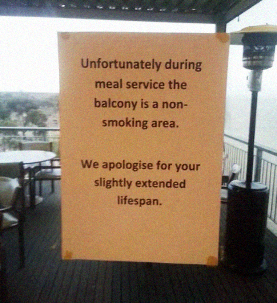 A sign on a balcony reads: "Unfortunately during meal service the balcony is a non-smoking area. We apologise for your slightly extended lifespan." Outdoor tables and chairs are in the background.