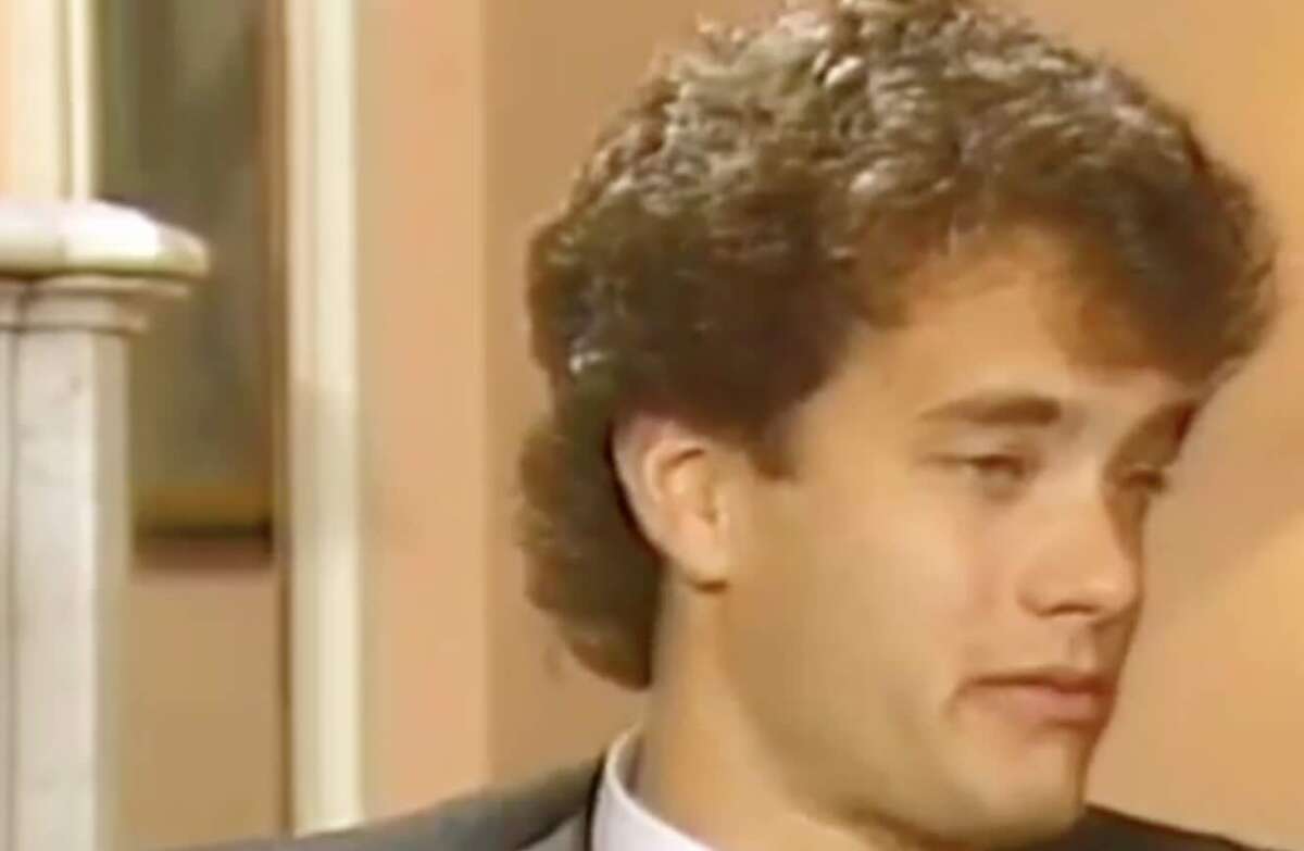 A young man with curly brown hair in a suit looks to the side in a softly lit indoor setting with beige walls.