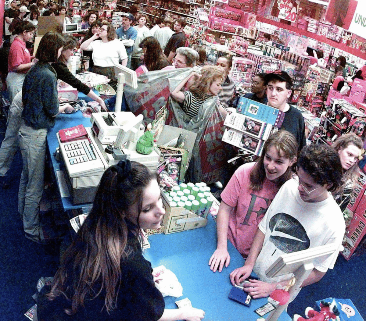 A crowded toy store with shoppers waiting in long lines at checkout counters. People hold boxes and shop bags, and shelves are filled with colorful toys and games. The atmosphere is busy and energetic.