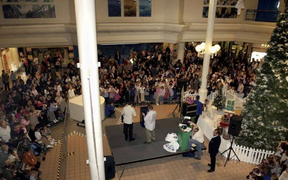 A large crowd gathers around a small stage in a shopping mall. Three people stand on the stage while others look on. There is a decorated Christmas tree and white picket fencing nearby. Shops and balconies surround the scene.