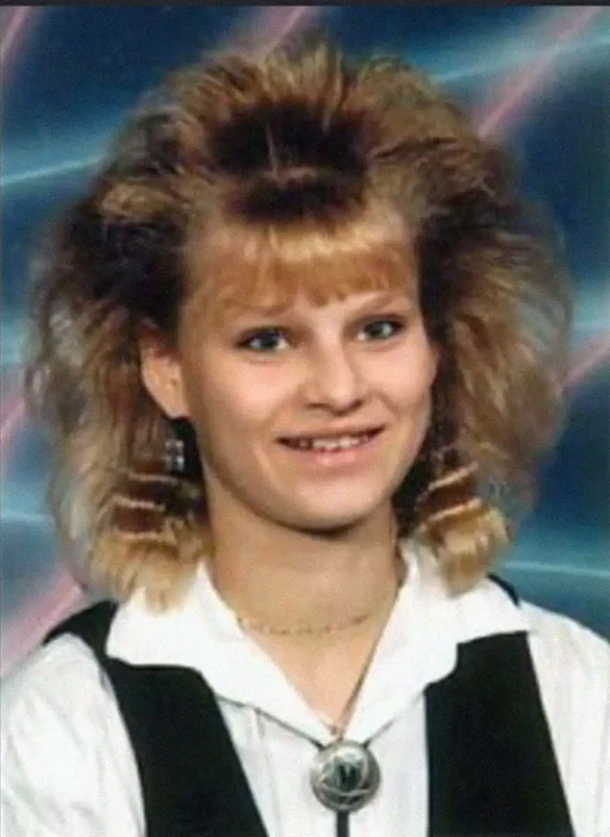 A young woman with blonde, teased, voluminous hair and bangs smiles at the camera. She wears a white shirt, a black vest, large earrings, and a bolo tie. The background features a retro laser pattern.