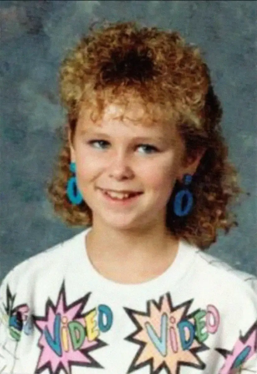 A young girl with curly, shoulder-length hair smiles slightly, wearing large blue earrings and a white shirt with colorful comic-style "VIDEO" text printed all over it against a mottled gray background.