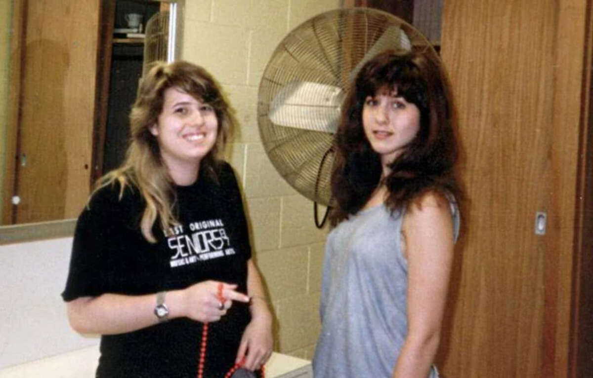 Two young women stand indoors; one wears a black T-shirt and smiles while holding a cord, the other wears a gray sleeveless top and looks at the camera. A large fan and a mirror are visible in the background.