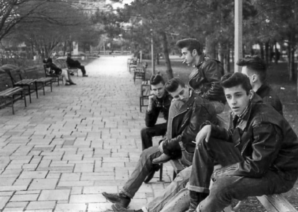 A group of young men in leather jackets sit on benches along a paved park path lined with trees, while other people sit further in the background. The image is black and white, evoking a vintage or retro style.