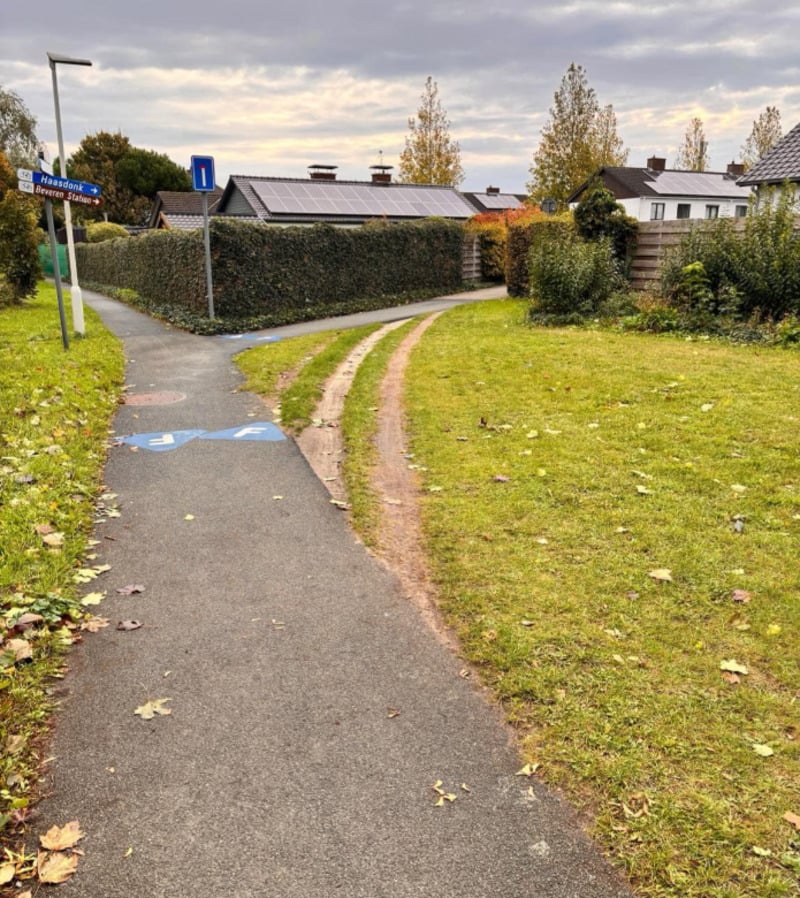 A paved pedestrian path with a street sign curves left, while a muddy shortcut cuts across the grass to the right. Houses and trees line the background under a cloudy sky.