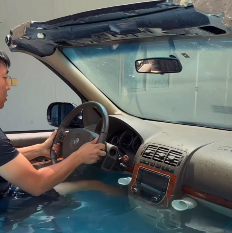 A person sits in the driver’s seat of a car with the roof removed, and water fills the interior up to the dashboard. The person grips the steering wheel, and sunlight shines through the windshield.
