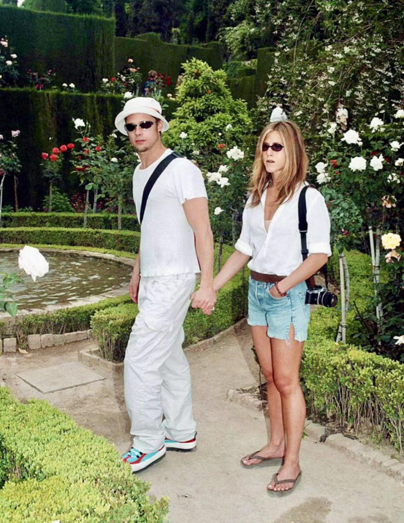 A man and woman wearing sunglasses hold hands while walking in a rose garden. Both are dressed casually in white tops; the man wears a hat and carries a bag, the woman has a camera over her shoulder and wears shorts and flip-flops.