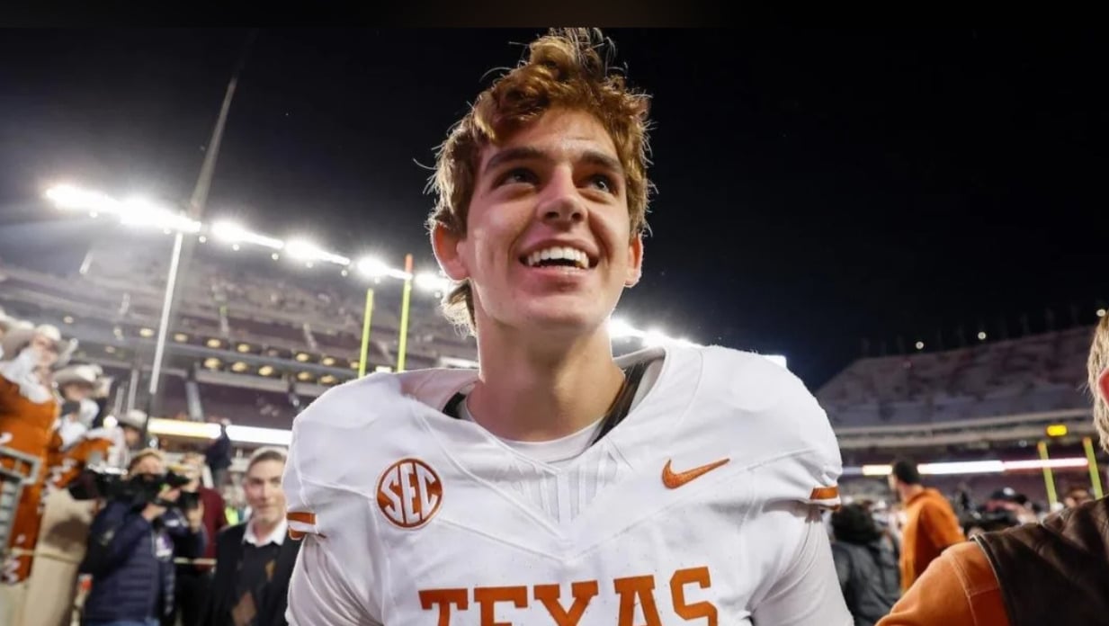 A smiling football player in a Texas Longhorns uniform stands on a football field at night, with stadium lights and a crowd in the background.