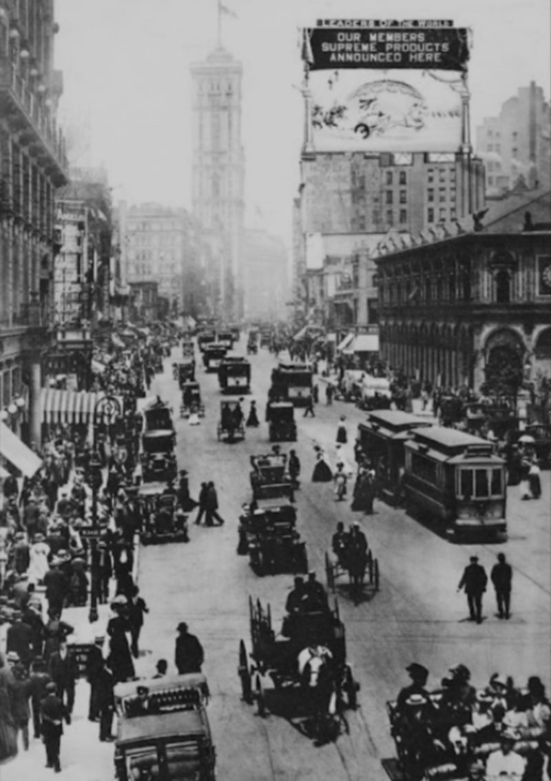 Historic black and white photo of a busy city street with cars, trolleys, and pedestrians; tall buildings line both sides and a large billboard is visible above the street.