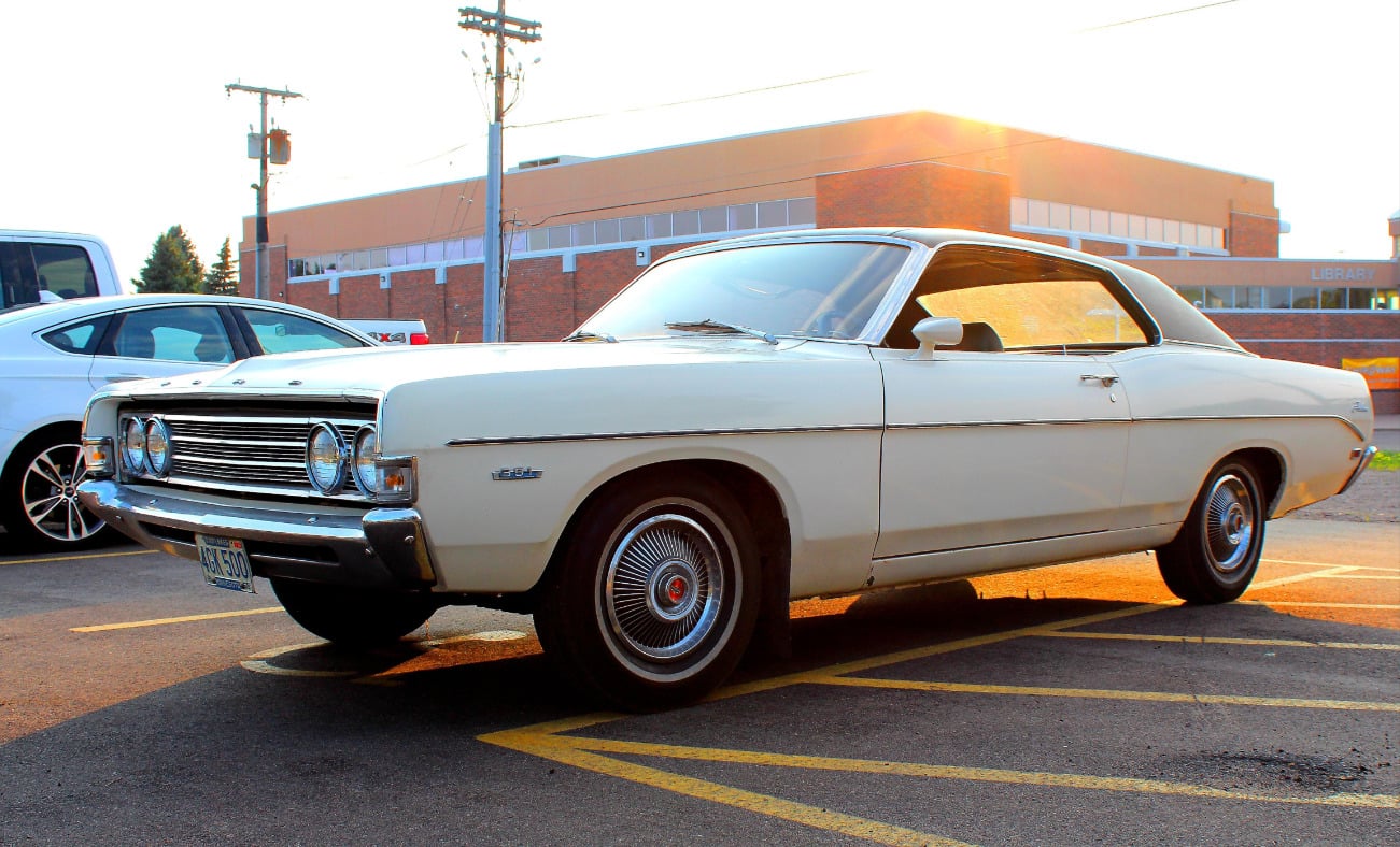 A vintage white two-door car is parked in a lot near a brick building at sunset, with sunlight reflecting off its surface and classic-style wheels visible.