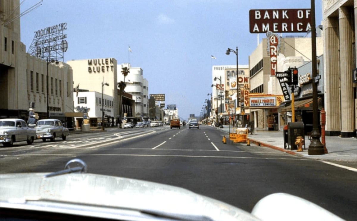 A wide street in a city with vintage cars, old storefronts, and large signs, including Bank of America and Mullen Bluett. The scene appears to be from the mid-20th century, with clear skies and light traffic.